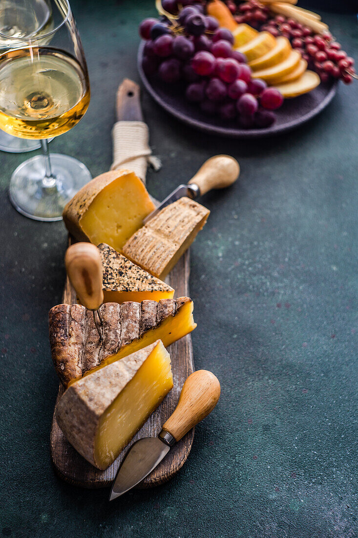 From above cheese variety on the wooden cutting board, fruit plate and wine glasses