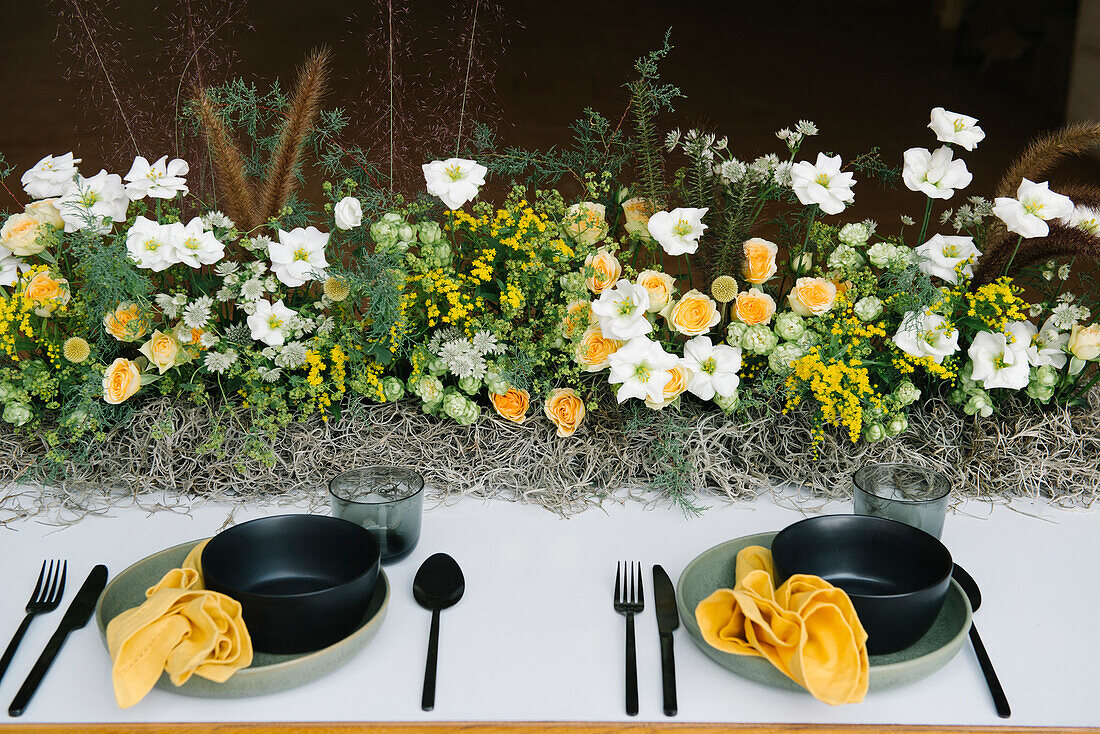 Blick von oben auf einen Teller und eine Schüssel mit Besteck und Serviette, die auf einem mit zarten frischen weißen und gelben Blumen dekorierten Tisch stehen