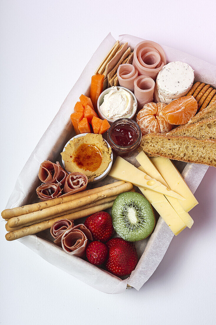 From above brunch box with assorted sliced meats various types of cheese and crispbreads arranged near ripe cup kiwi sweet strawberries and peeled mandarin near jam in glass jar on white background