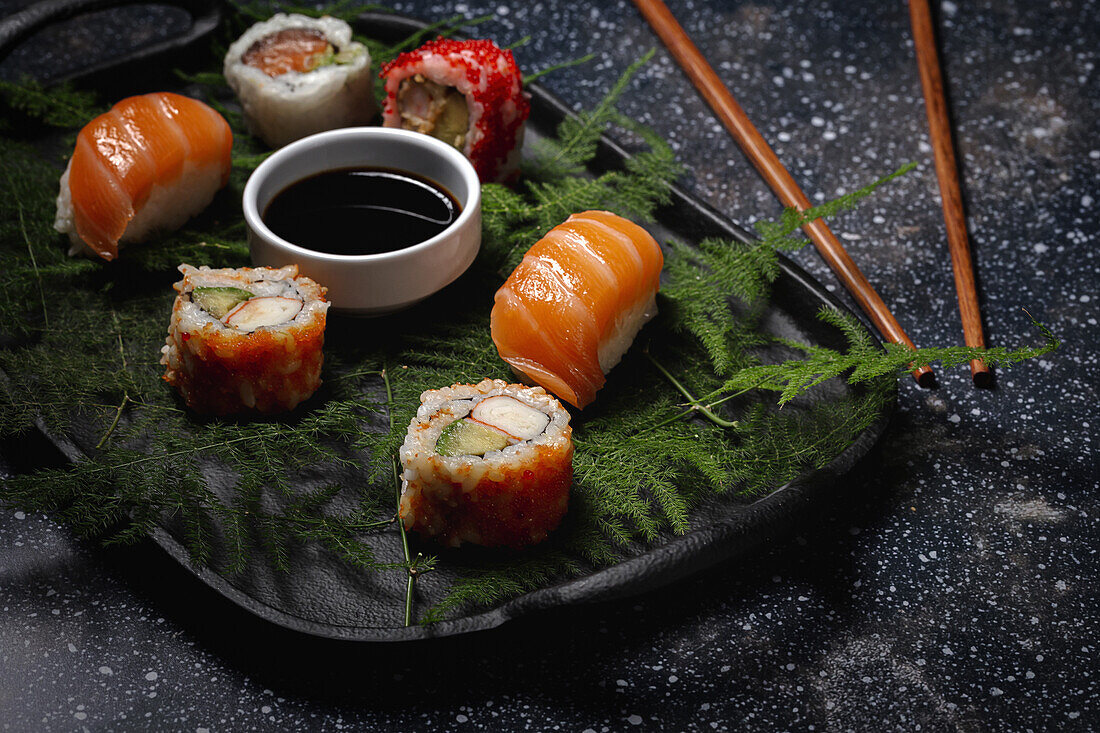 Tasty fresh assorted sushi served on green plant twigs on black plate with soy sauce on marble table near chopsticks