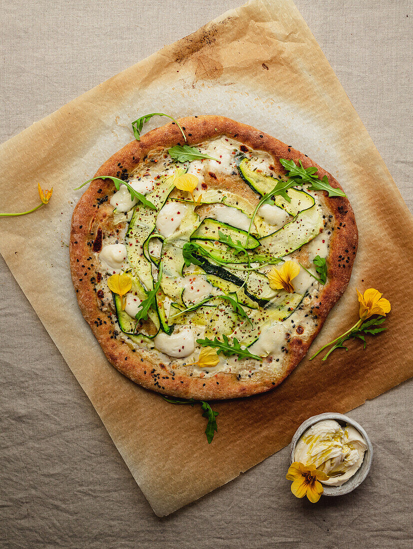 Top view of tasty pizza with squash slices and condiments with fresh arugula leaves on parchment paper on beige background
