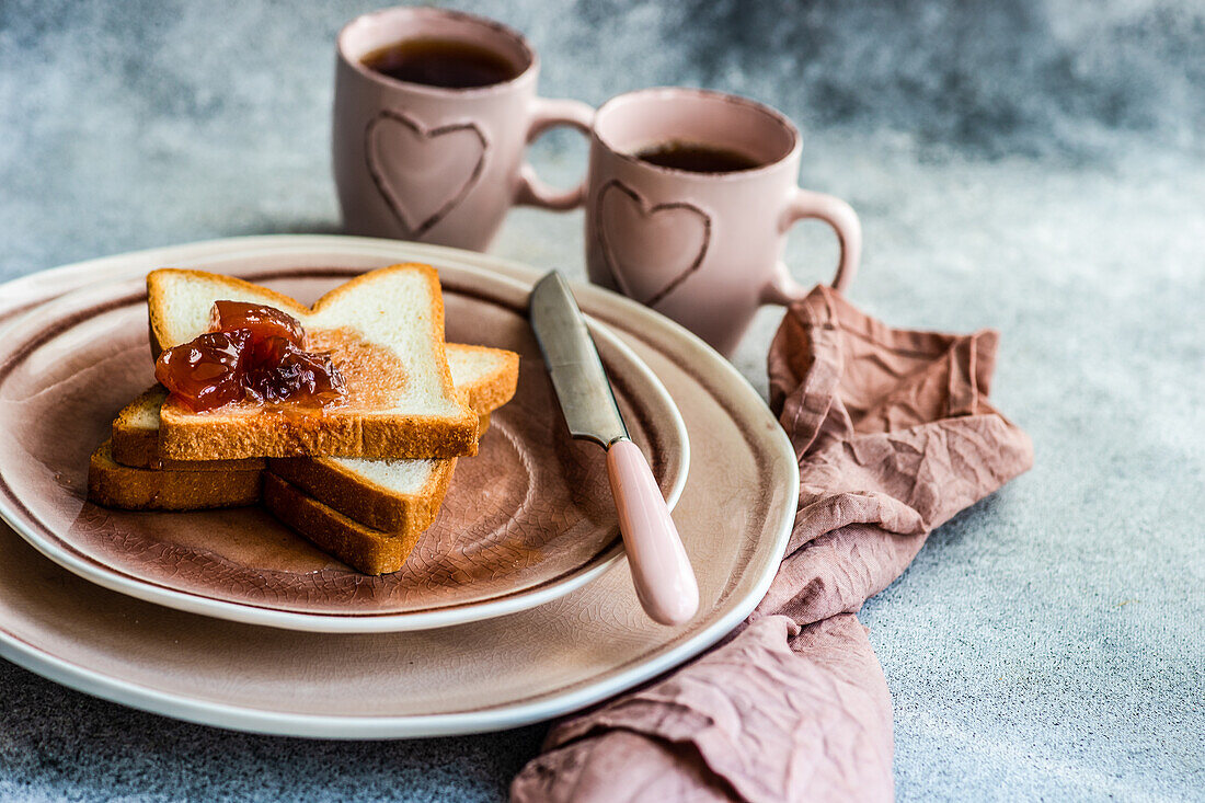Traditionelles Frühstück mit Marmelade auf Toasts in rosa Keramiktellern und einer Tasse Tee auf dem Betontisch