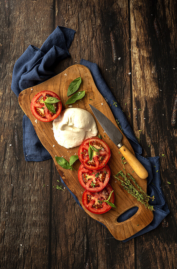 Draufsicht auf köstlichen Mozzarella und Scheiben frischer Tomaten mit grünen Basilikumblättern und Thymian, serviert auf einem hölzernen Schneidebrett mit Messer auf dem Tisch