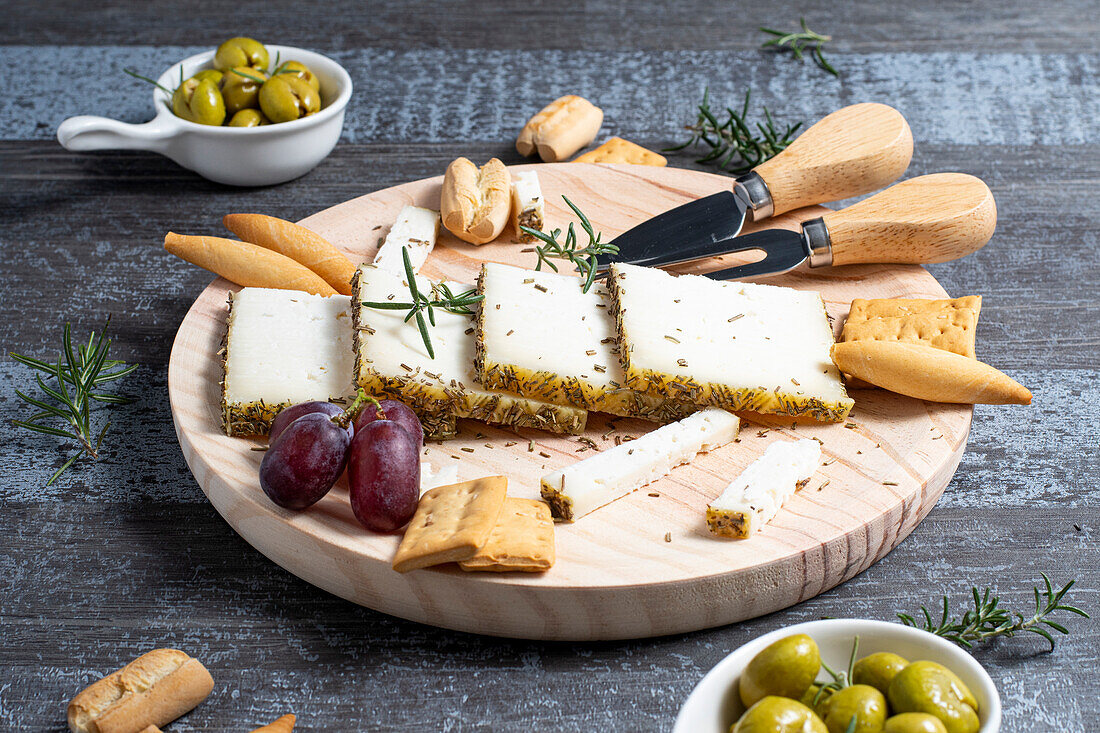 Appetizing cheese served on wooden table with ripe grapes and crackers decorated by rosemary sprigs near olives in bowls on table