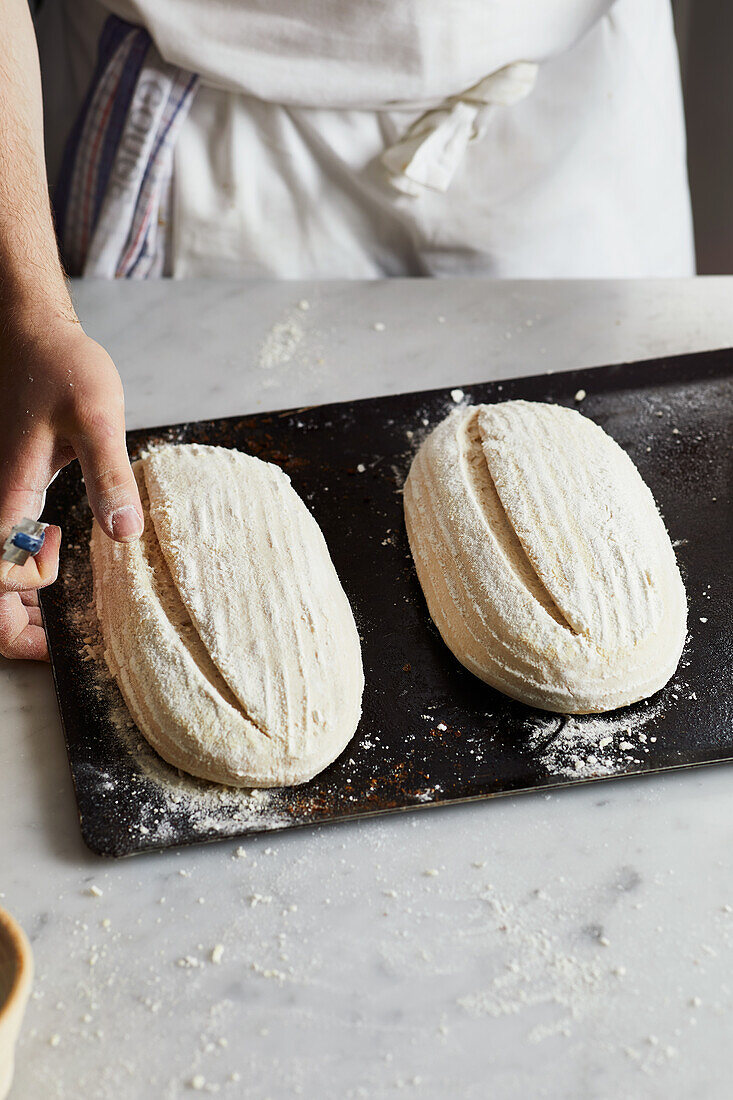 Anonymer Bäcker in Schürze mit Klinge ritzt rohen Sauerteig Laib auf Tisch während der Vorbereitung Brot in der Küche