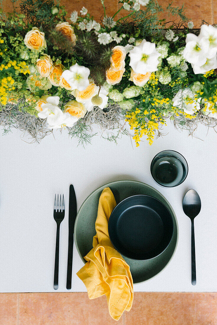 Draufsicht auf Teller und Schüssel mit Besteck und Serviette, die auf einem mit zarten frischen weißen und gelben Blumen geschmückten Tisch serviert werden