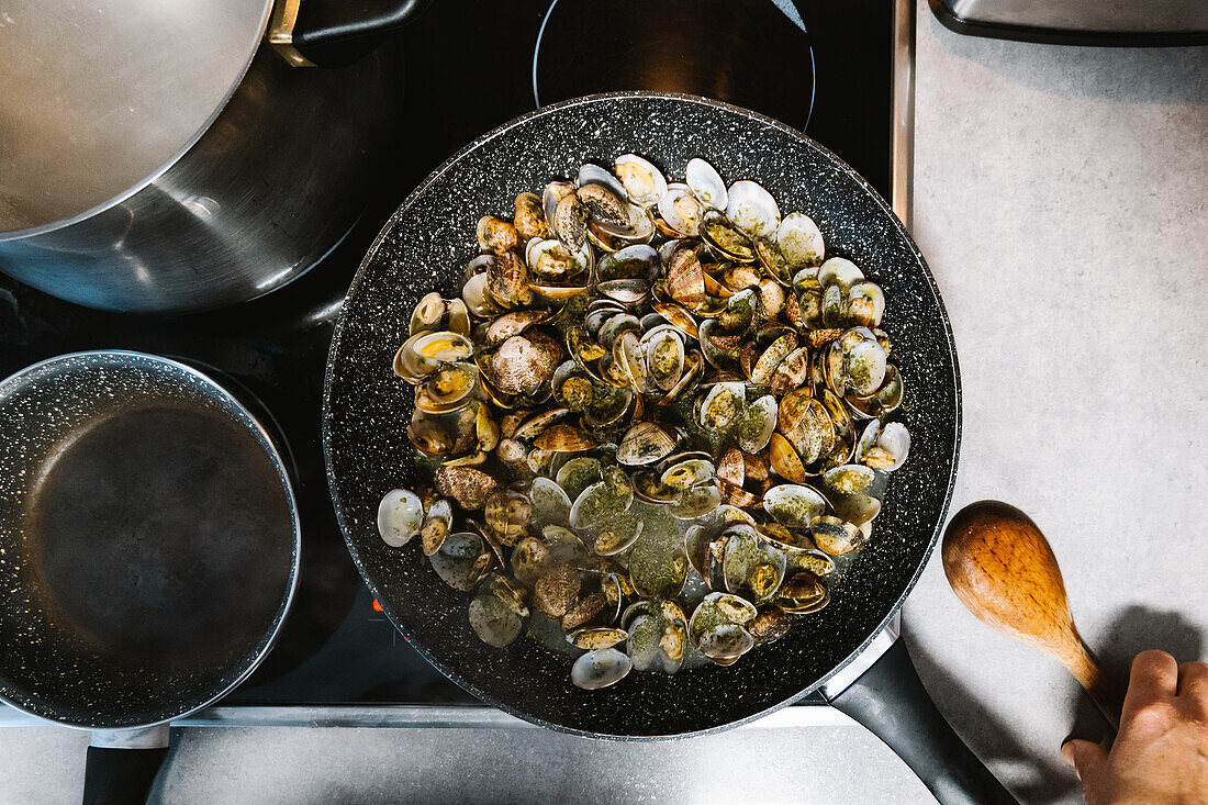 Haufen schmackhafter Mollusken mit Schalen, die in einer heißen schwarzen Bratpfanne auf einem Elektroherd in einer hellen Küche zu Hause gebraten werden