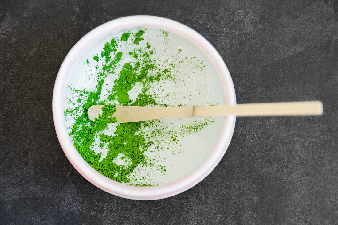 From above wooden chashaku of dried green tea adding to ceramic bowl placed on table against grey background