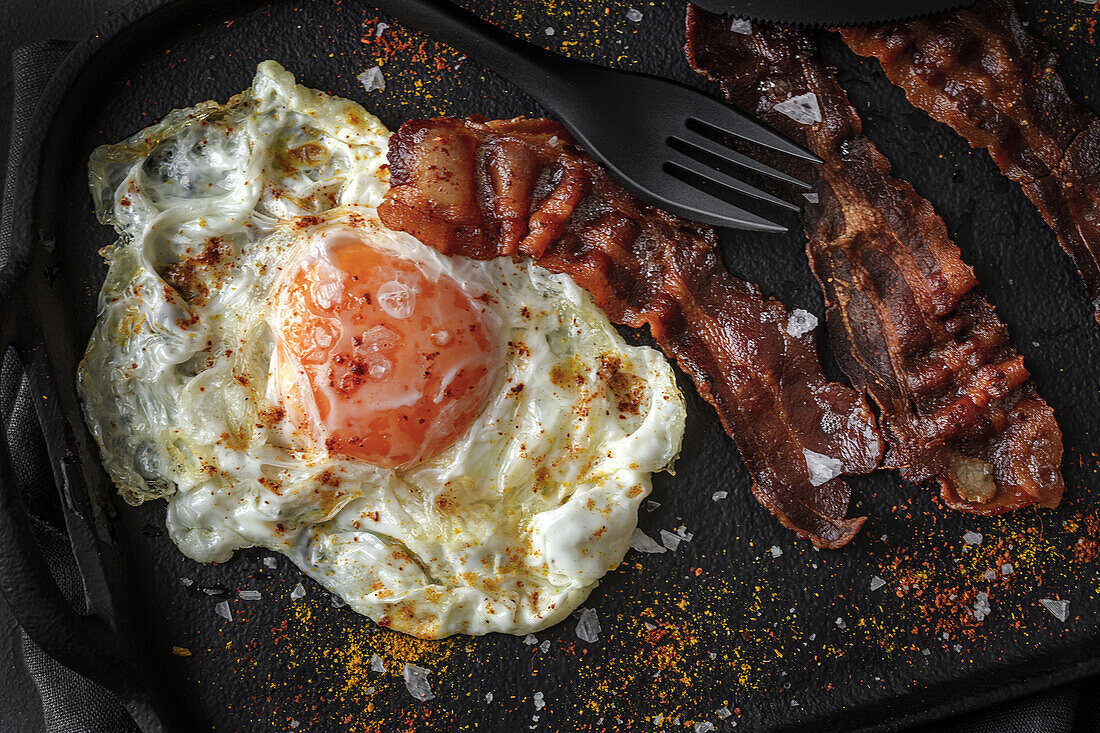 Top view of sunny side up egg with fried bacon slices and condiments on tray against cutlery on dark background