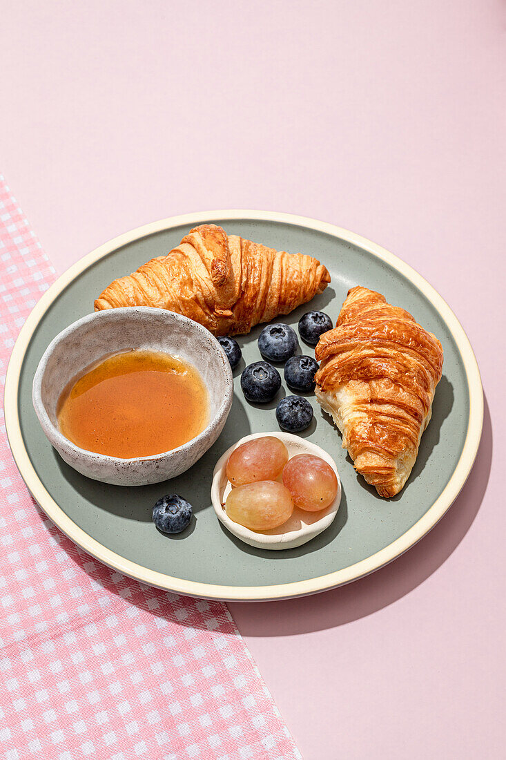 Leckere Croissants auf einem Keramikteller mit frischen Blaubeeren und Marmelade auf einem rosa Tisch