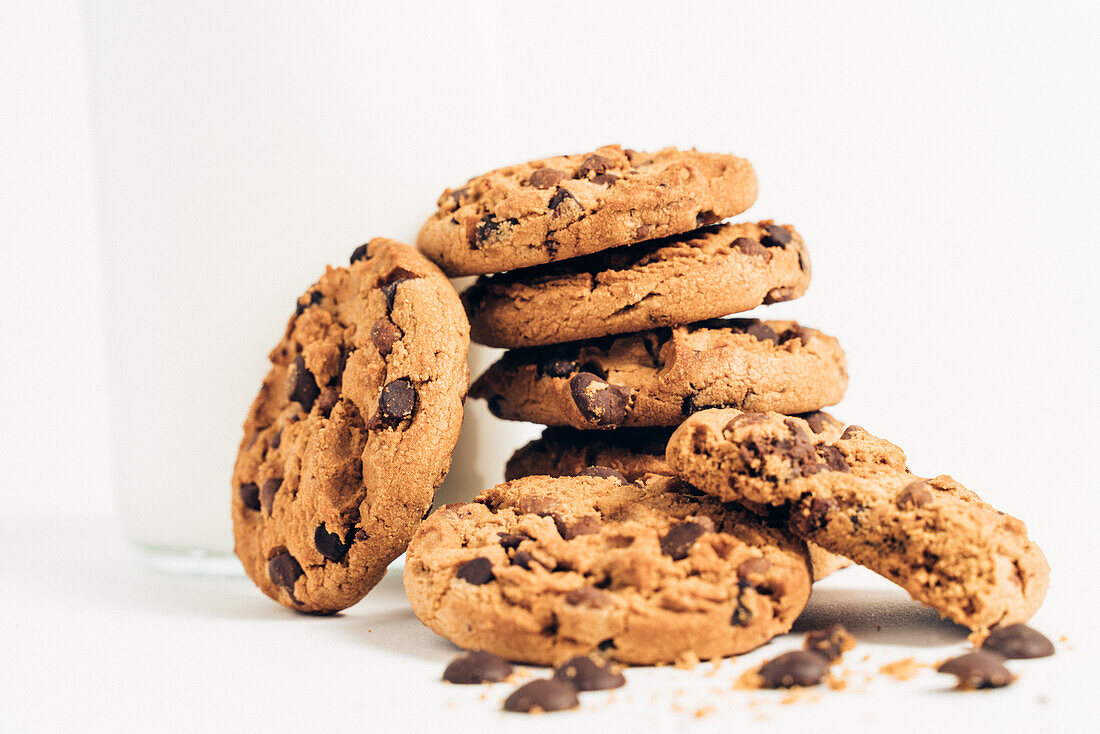 Pile of sweet crunchy cookies with chocolate chips placed on table with crumbs and milk on white background in room