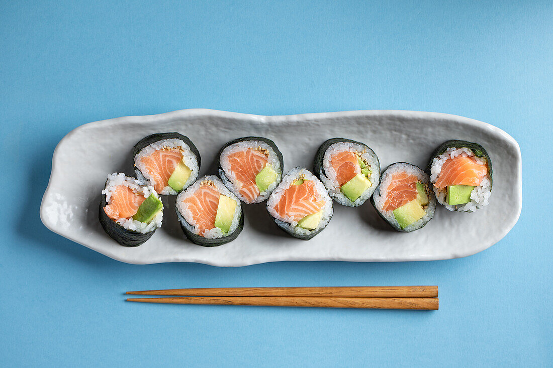 From above set of tasty Futomaki Norwegian sushi rolls with salmon and avocado served near chopsticks on blue background in light studio