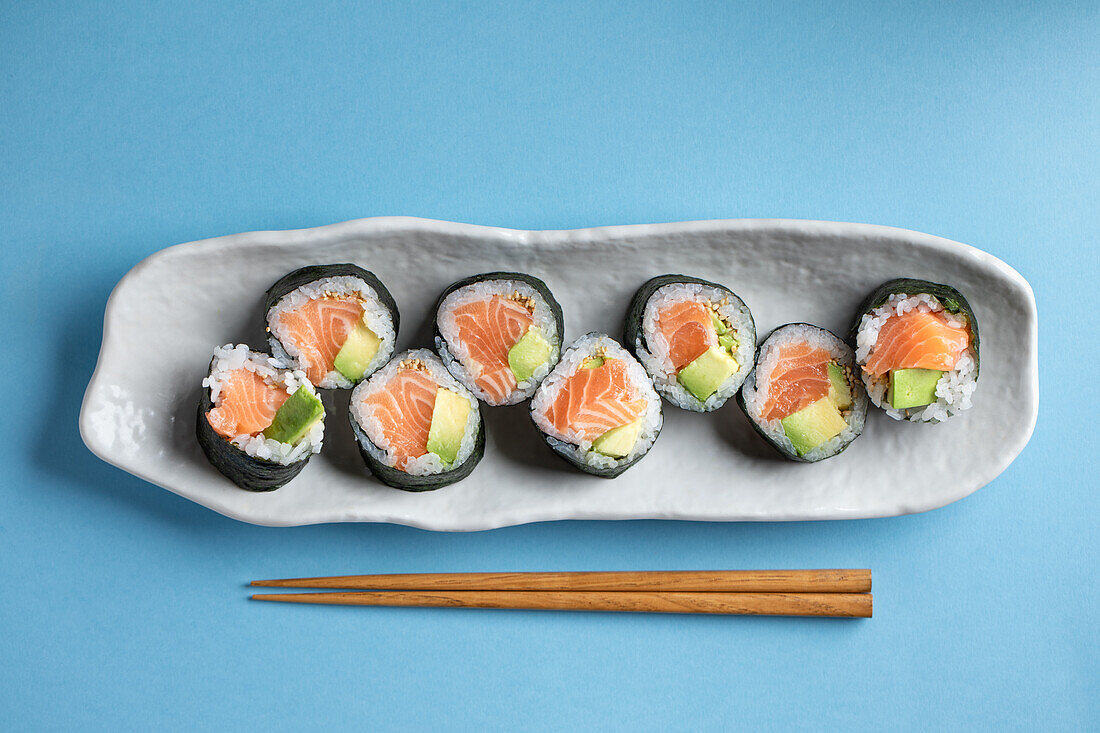Leckere norwegische Futomaki-Sushi-Rollen mit Lachs und Avocado, serviert mit Stäbchen auf blauem Hintergrund in einem hellen Studio