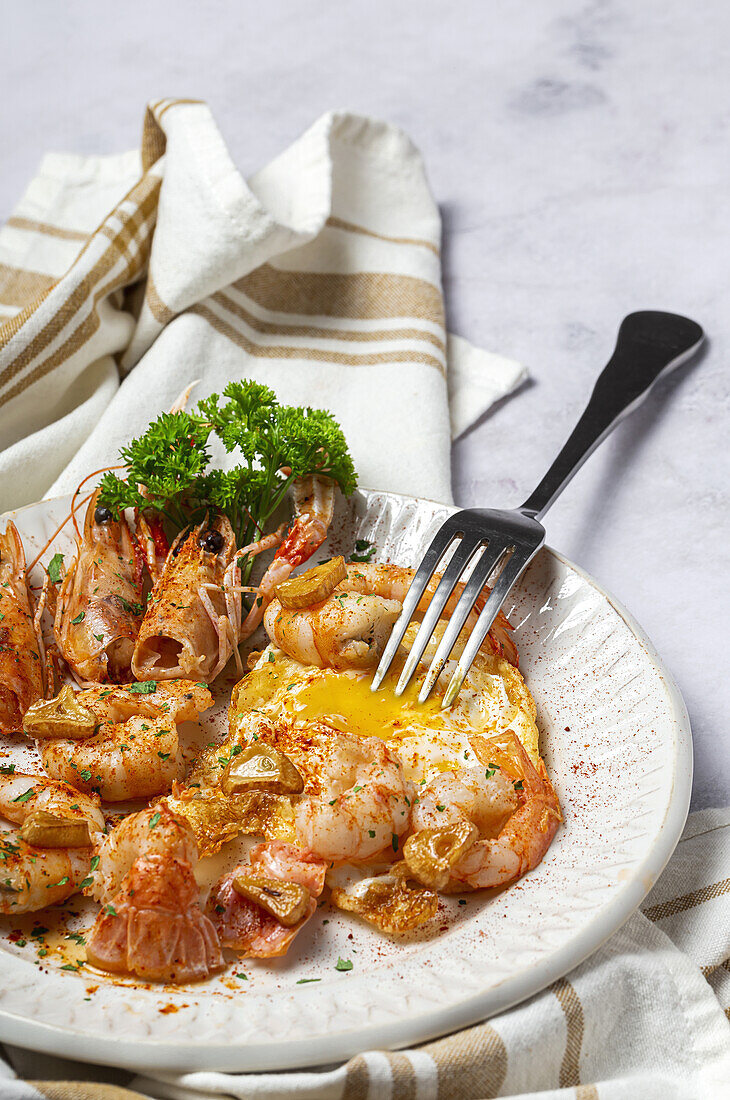 From above appetizing fried eggs with king prawns served on white plate with green herbs and seasoning on towel in light kitchen