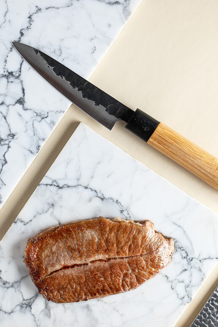 Draufsicht auf schmackhaftes Kobe-Präfektur-Wagyu-Rindfleisch, serviert auf einem Marmorschneidebrett neben einem scharfen Messer in einer hellen Küche während des Kochvorgangs