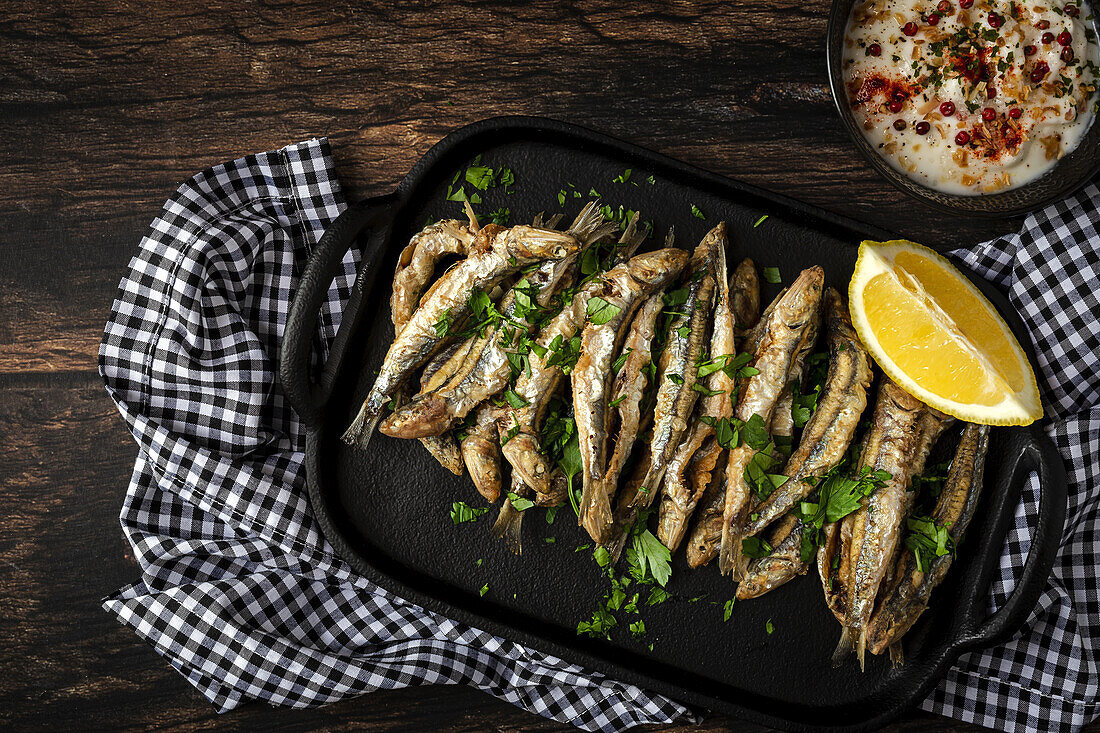 Top view of tasty fried anchovies with juicy lemon piece and chopped parsley on tray against savory sauce