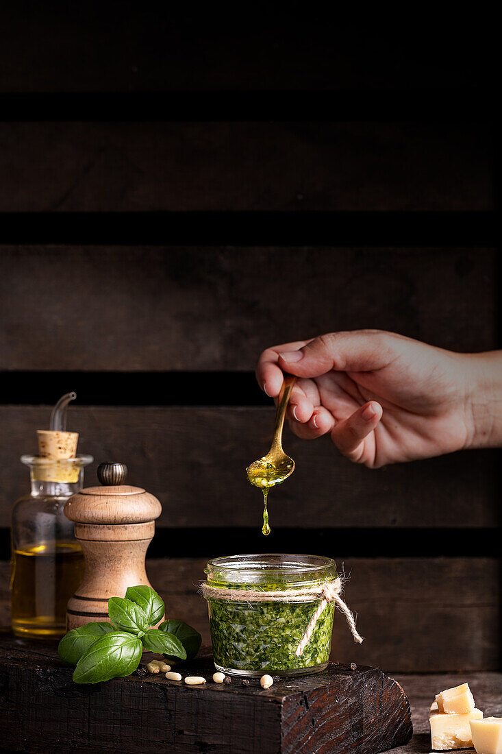 Anonyme Person mit Löffel, die Olivenöl in einen Topf mit frisch zubereiteter Pestosauce gibt