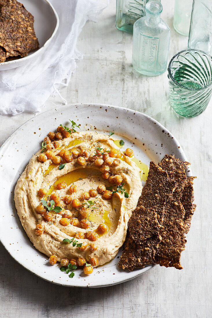 Draufsicht auf köstlichen Hummus mit Kichererbsensamen und Olivenöl, serviert auf einem Teller mit knusprigen gesunden Crackern auf einem Tisch in der Küche