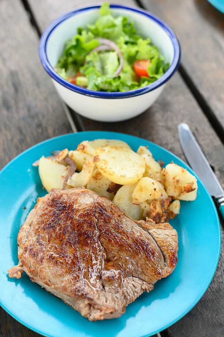 Draufsicht auf ein appetitliches gebratenes Steak mit Kartoffeln, serviert auf einem blauen Teller mit Gabel und Messer neben einer Schüssel mit Salat auf einem Holztisch