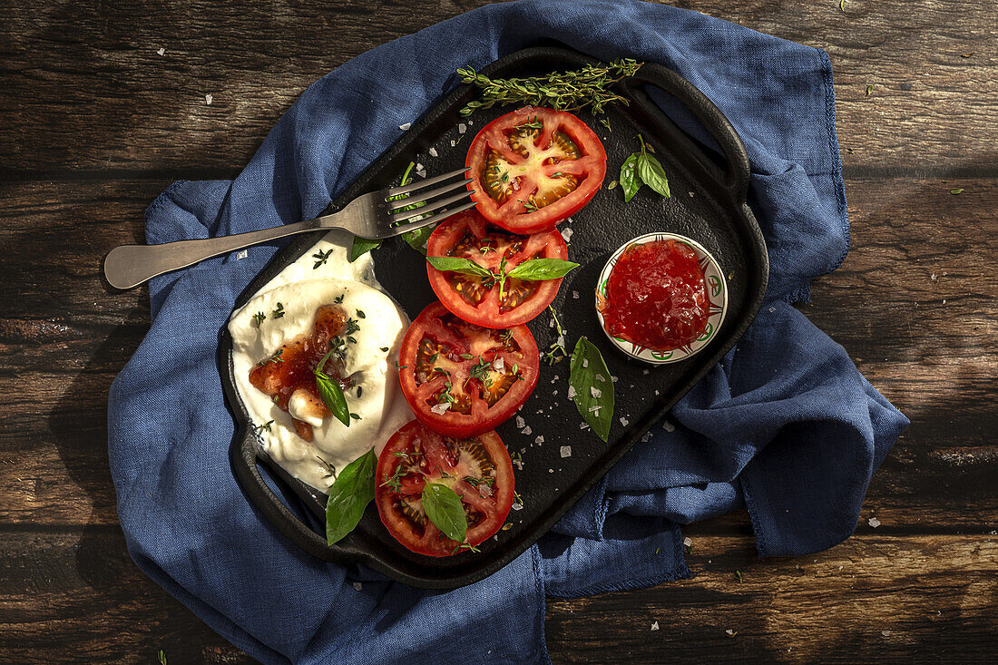 From above of appetizing burrata cheese with sauce and slices of fresh tomato with thyme and basil leaves served on cast iron tray placed on table cloth on wooden table with fork