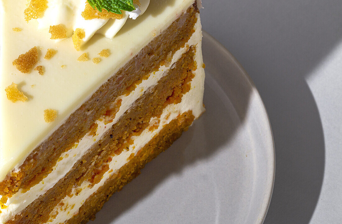 From above close up of slice of tasty sweet carrot sponge cake with cream decorated with mint leaf served on plate with spoon on table on white background