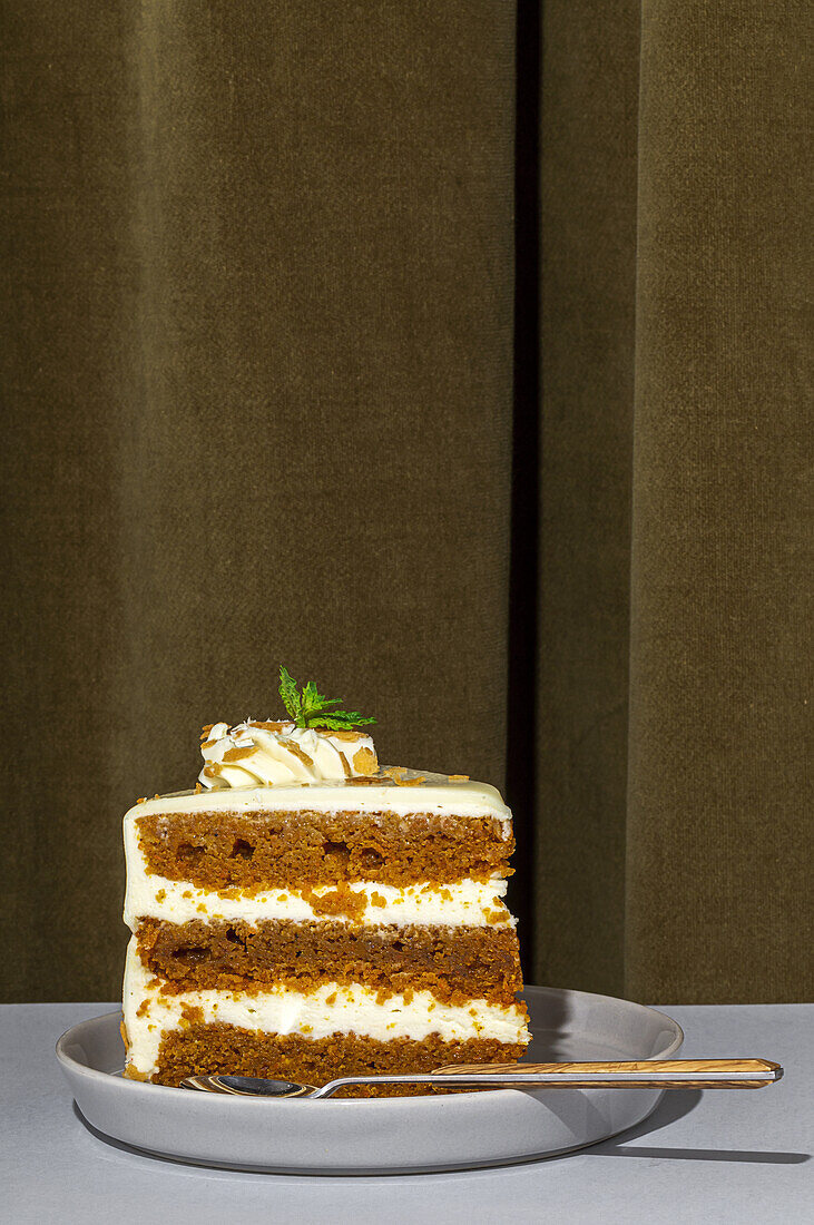 Slice of tasty sweet carrot sponge cake with cream decorated with mint leaf served on plate with spoon on table in light room with curtain on the background