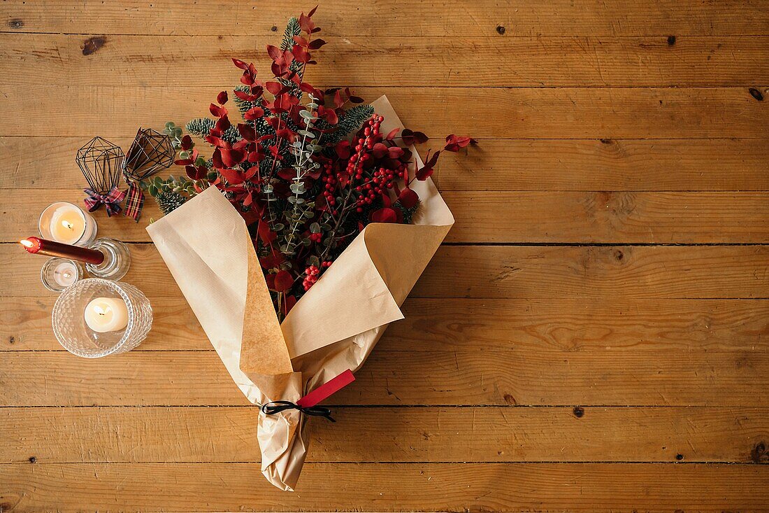 Festlicher, stilvoller, dekorativer Weihnachtsstrauß mit Eukalyptuszweigen und leuchtend roten Zweigen mit Beeren auf einem Holztisch mit Kerzen im Raum von oben