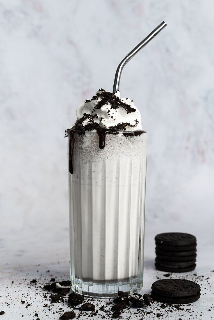 Glass of white sweet milkshake topped with whipped cream and chocolate cookie crumbs served with straw on messy table near wall