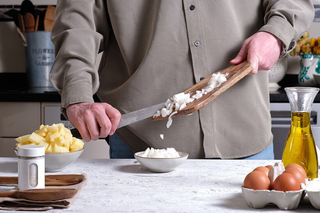 Unbekannte Person legt gehackte Zwiebeln vom Schneidebrett in eine Schüssel, während sie Zutaten für ein spanisches Omelett oder eine Tortilla mit Kartoffeln in einer Küche zu Hause vorbereitet