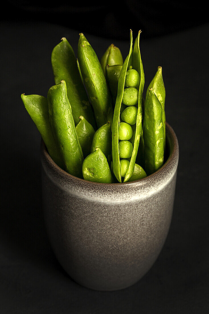 Bunch of ripe green pods with fresh peas placed in ceramic pot against black background