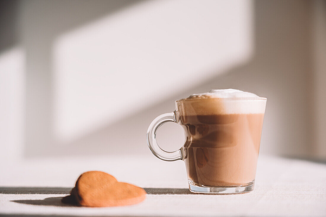 Glass of foamy latte macchiato served on table with heart shaped cookies on sunny day in kitchen