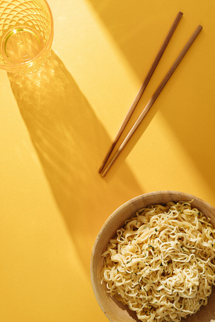 Top view bowl of delicious noodles with seasoning placed on yellow background with wooden chopsticks and glass in light room