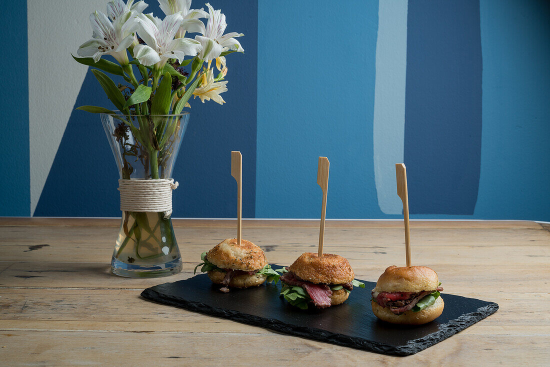 Fresh appetizing mini burgers served on black board near vase with blooming lilies against blue wall