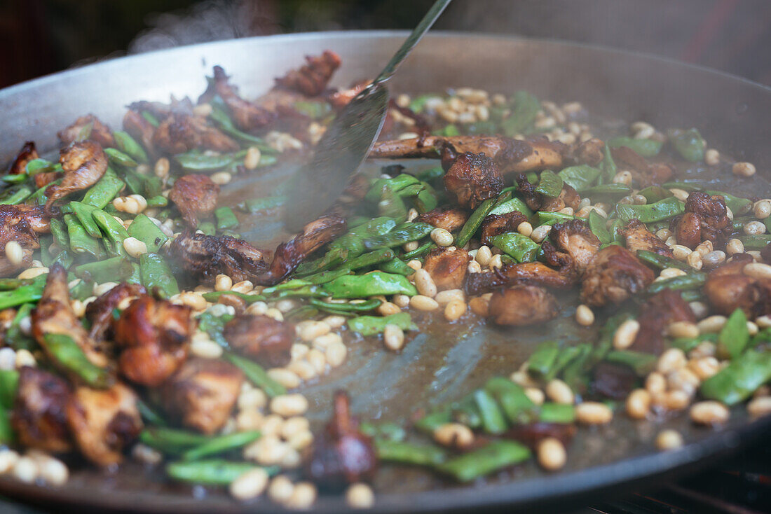 Nahaufnahme eines nicht erkennbaren Kochs, der ein appetitliches Gericht mit Huhn und grünen und weißen Bohnen in einer Paellapfanne rührt