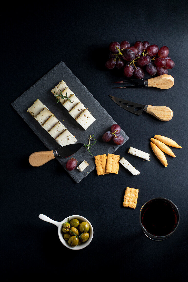Ansicht von oben auf ein schwarzes Brett mit verschiedenen Käsesorten, reifen Weintrauben und Crackern, die neben Messern auf einer dunklen Fläche liegen