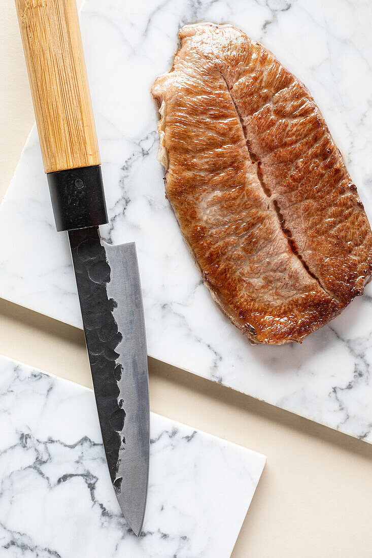 Top view of tasty kobe prefecture wagyu beef served on marble cutting board near sharp knife in light kitchen during cooking process