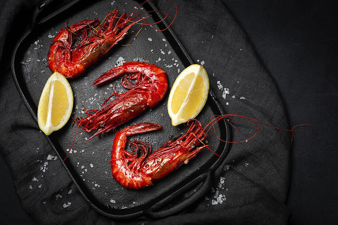 Leckere gekochte rote Garnelen auf einem Tablett mit grobem Salz und saftigen Zitronenstücken auf dunklem Hintergrund