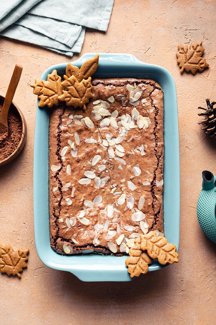 Top view of tasty baked brownie with almond flakes in baking tray with cookies placed on table with cocoa powder