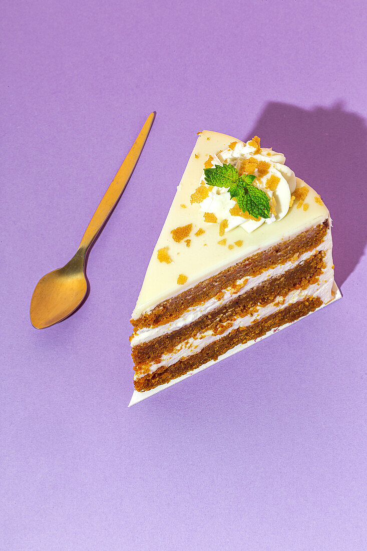 From above close up of slice of tasty sweet carrot sponge cake with cream decorated with mint leaf served on plate with spoon on table on purple background