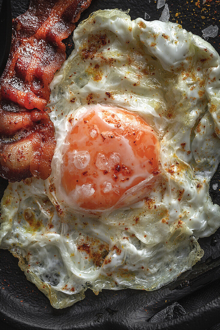 From above of sunny side up egg with fried bacon slices and condiments on dark tray