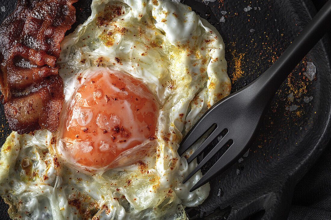 From above of sunny side up egg with fried bacon slices and condiments on tray against cutlery on dark background