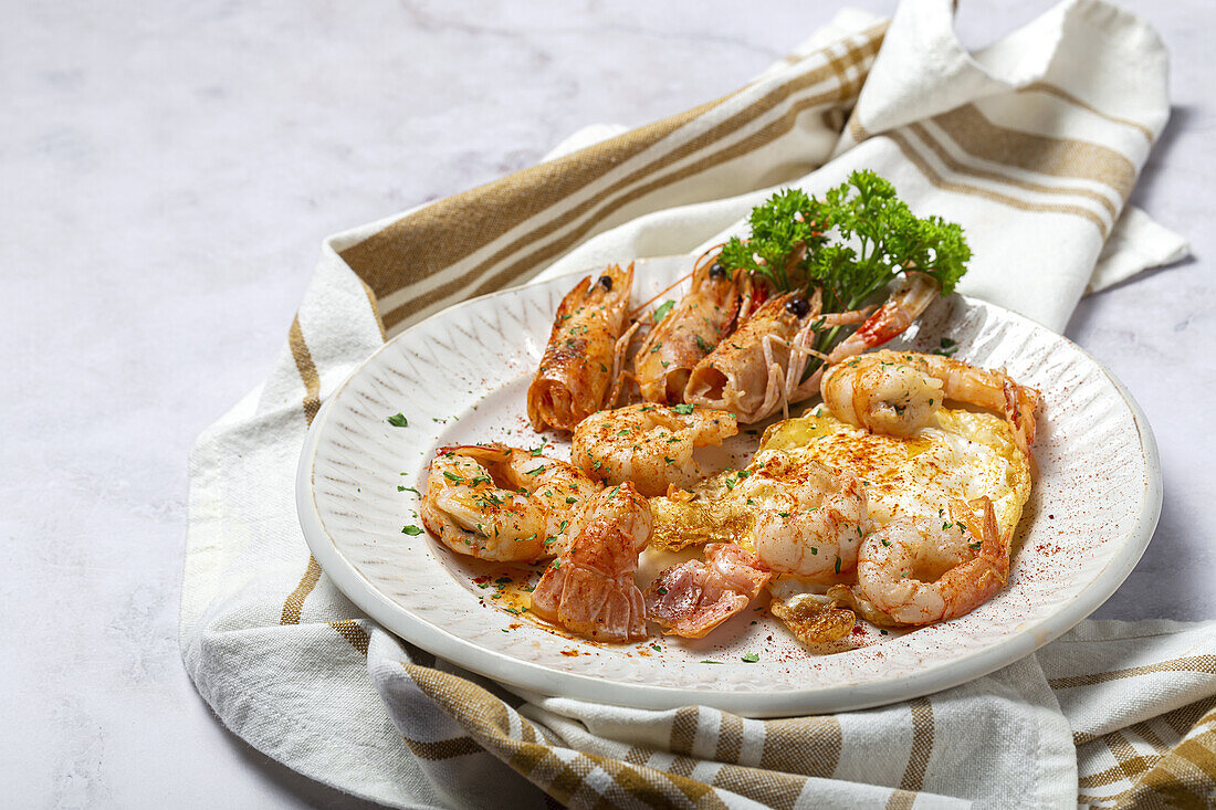 Appetitliche Spiegeleier mit Riesengarnelen auf weißem Teller mit grünen Kräutern und Gewürzen auf einem Handtuch in einer hellen Küche von oben