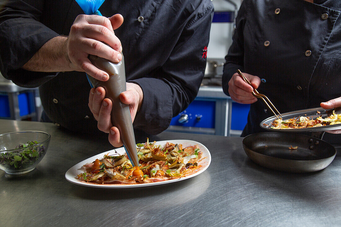 Männlicher Koch drückt Soße aus einer Tüte auf einen Teller mit einem leckeren Gericht, während er in der Nähe eines Kollegen steht und in einer Restaurantküche arbeitet