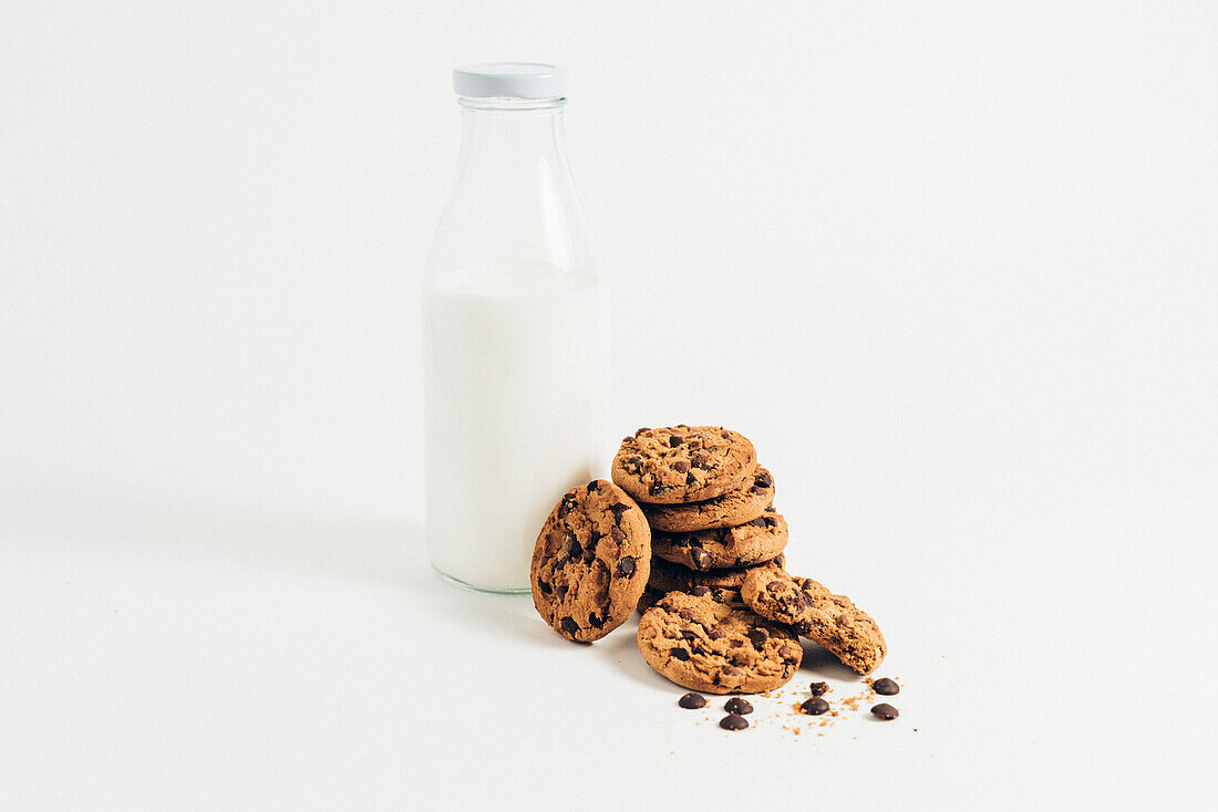Pile of sweet crunchy cookies with chocolate chips placed on table with crumbs and milk on white background in room