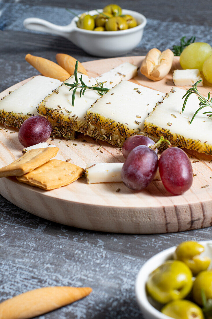 Appetizing cheese served on wooden table with ripe grapes and crackers decorated by rosemary sprigs near olives in bowls on table
