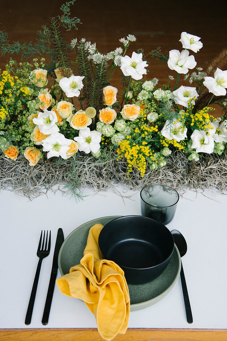 Blick von oben auf einen Teller und eine Schüssel mit Besteck und Serviette, die auf einem mit zarten frischen weißen und gelben Blumen dekorierten Tisch stehen