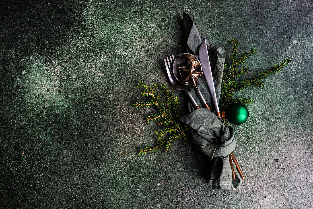 Von oben gedeckter Tisch für ein Weihnachtsessen mit dunklem, modernem Besteck, eingewickelt in eine grüne Serviette mit Tannenbaumblättern und Dekor auf einem Betontisch