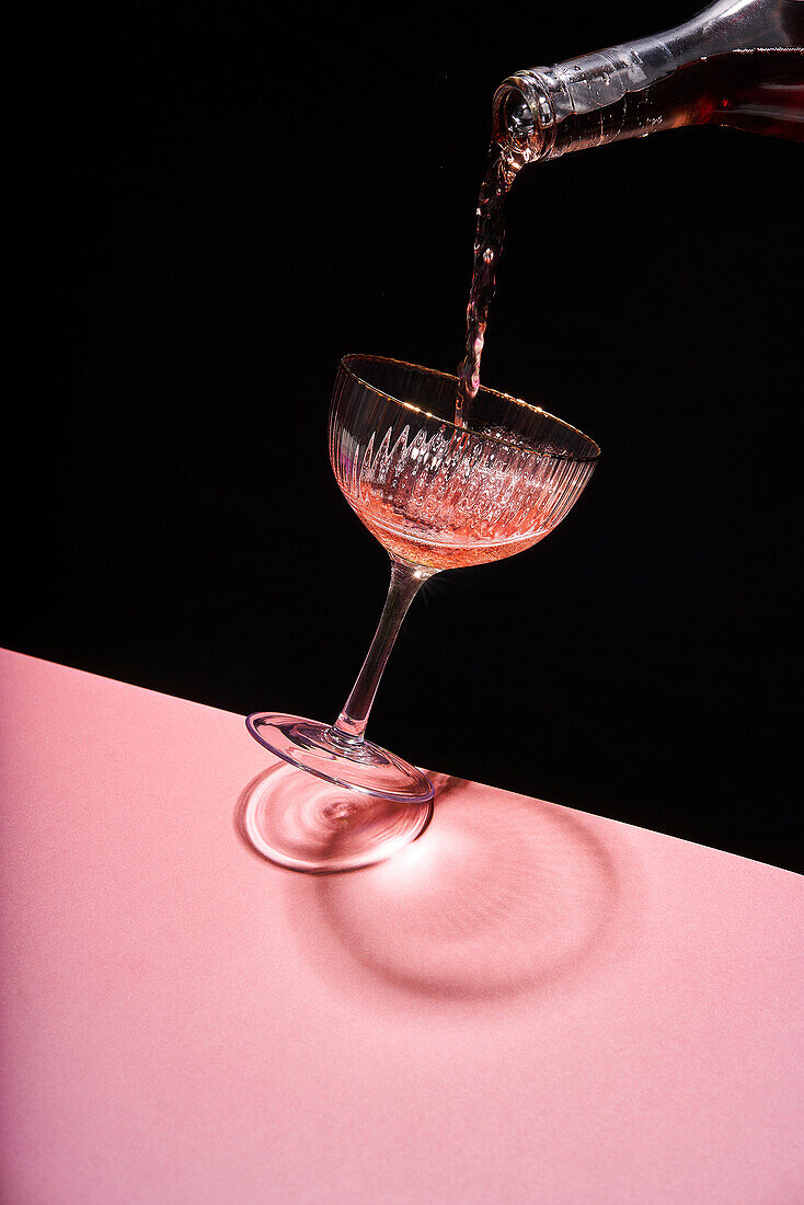 Anonyme Person gießt Rosensekt in ein elegantes Coupe-Glas vor zweifarbigem Hintergrund
