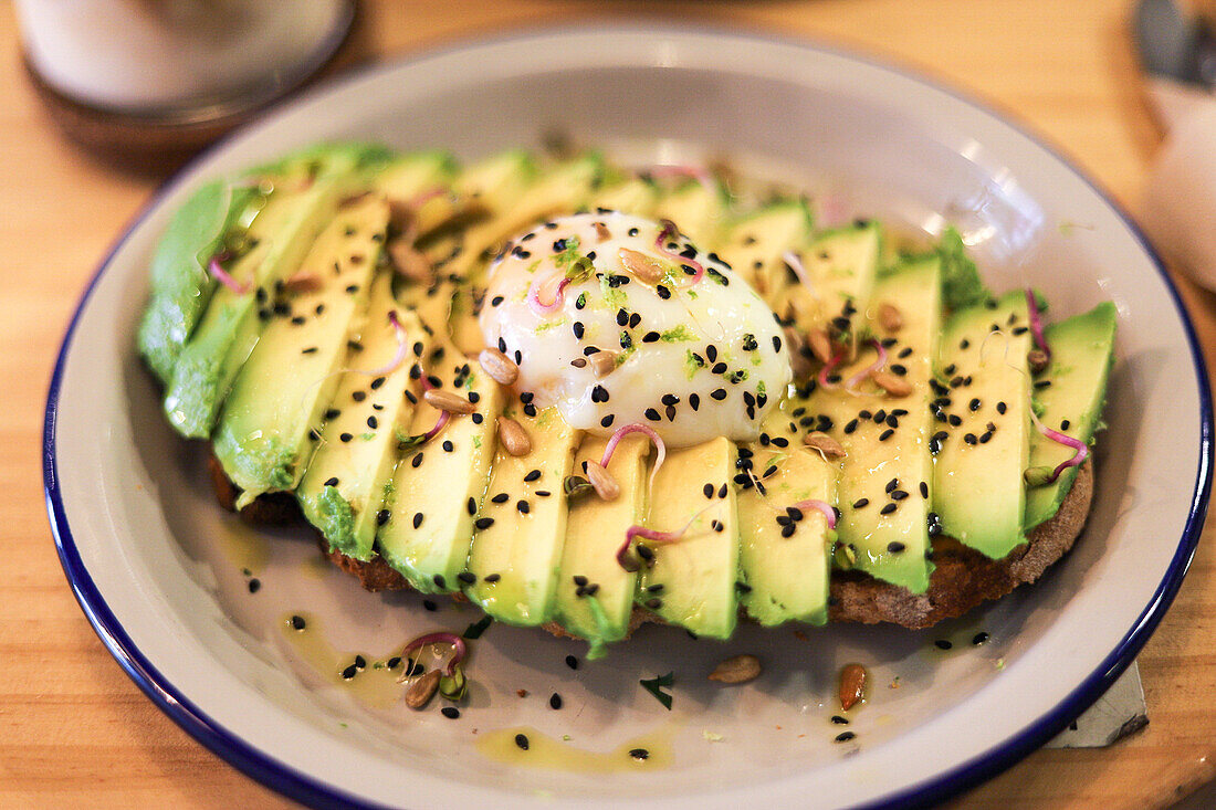 From above of appetizing toast with sliced avocado and poached egg sprinkled with chia seeds served on ceramic plate placed on wooden table