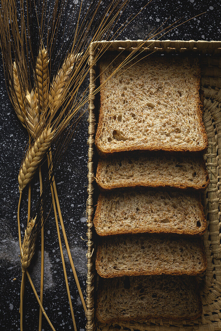 Top view of fresh homemade rye bread near knife in wicker basket and wheat spikes on table